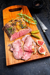 Traditional lunch meat with sliced cold cuts roast beef with gherkin and fig fruit as closeup on a modern design cutting board