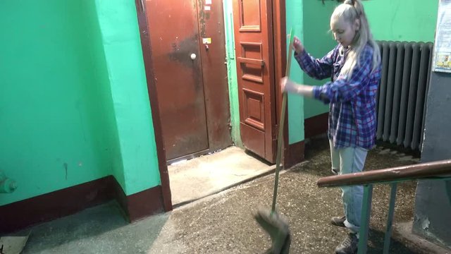 A young woman in blue jeans and a cage shirt swings and washes the floor on a staircase next to an elevator in a Stalinist-built apartment building, Cleaning public spaces by a labor immigrant cleane