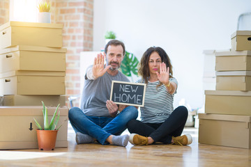 Fototapeta na wymiar Middle age senior couple sitting on the floor holding blackboard moving to a new home with open hand doing stop sign with serious and confident expression, defense gesture