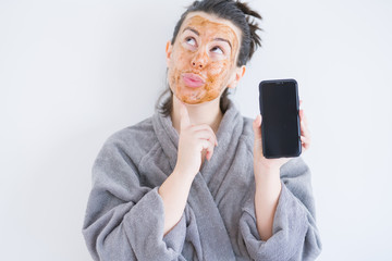 Beautiful woman wearing cosmetic facial mask as skincare treatment showing smartphone screen...