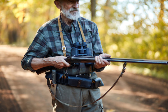 Older hunter man with grey beard holding gun to hunt on birds, ready to shoot. Forest background