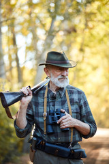 Portrait of senior bearded huner holding shotgun, using binocular while hunting on wild animals, look away