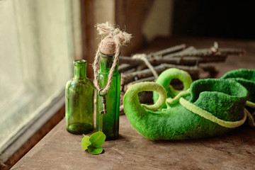 Colorful felted green shoes stand next to antique glass bottles, a leaf of clover and a bundle of firewood