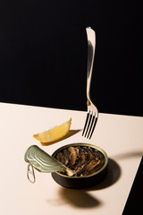 Opened tin fish, sardine with a slice of lemon on powder colored table with black background. A fork is floating above. Abstract conception.