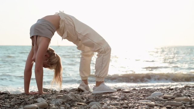 A lithe athletic woman by the sea leans back arching her back to put her hands on the floor while exercising to maintain mobility