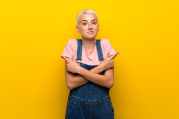 Teenager girl with overalls on yellow background pointing to the laterals having doubts