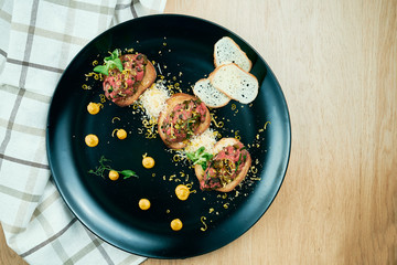 Beef tartare on fried croutons served on a stylish black plate. Restaurant Food. Wooden bakcground. Close up