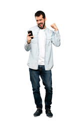 Full-length shot of Handsome man with beard with phone in victory position over isolated white background