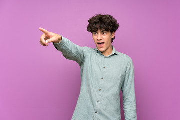 Young man over isolated purple wall pointing away