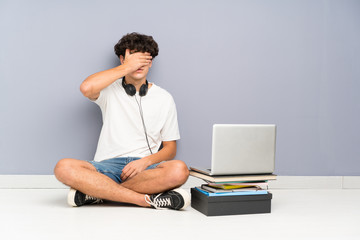 Young man with his laptop sitting one the floor covering eyes by hands