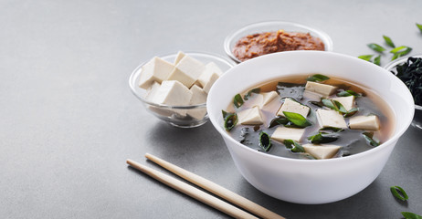 Bowl with miso soup, wakame seaweed, miso pasta, tofu and chopsticks on a gray background.