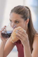 Pretty girl drinking soda which is served in a glass cup which she holds with one of her hands while with the other hand she holds a piece of bread.