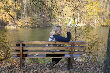 A girl sits on a bench against the background of a lake and autumn forest and drinks coffee in a glass. The girl is resting on a bench in the autumn forest in front of the lake and drinking coffee.