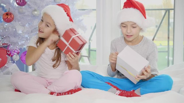 Full-body video of cute children sitting in lotos position with Christmas presents, shaking, knocking, listening to what is inside.