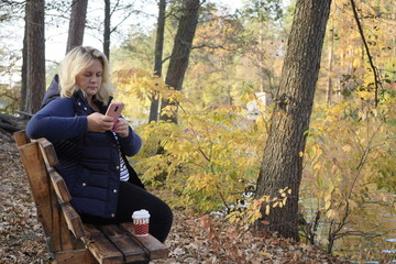 Fototapeta premium Girl sitting on a bench uses a smartphone on the background of the lake and autumn forest. A girl sits on a bench in the autumn forest in front of the lake.