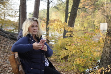 Girl sitting on a bench uses a smartphone on the background of the lake and autumn forest. A girl sits on a bench in the autumn forest in front of the lake.