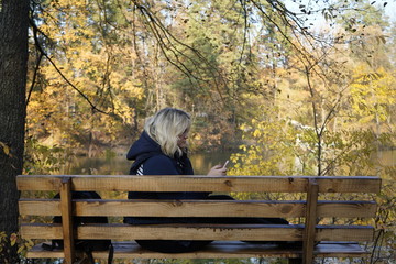 Girl sitting on a bench uses a smartphone on the background of the lake and autumn forest. A girl sits on a bench in the autumn forest in front of the lake.