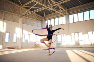 Active beautiful rhythmic gymnast standing in Sur le cou-de-pied pose, waving long colourful ribbon...