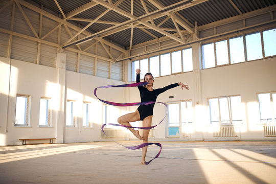 Active beautiful rhythmic gymnast standing in Sur le cou-de-pied pose, waving long colourful ribbon in the air, dancing in bright sports hall, professional sport concept