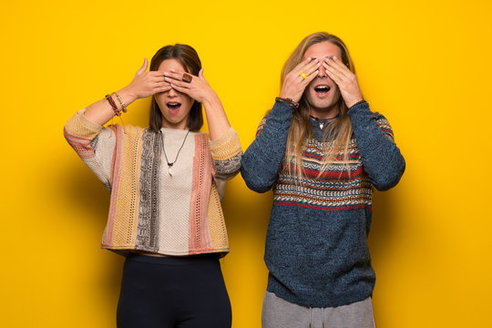 Hippie couple over yellow background covering eyes by hands. Surprised to see what is ahead