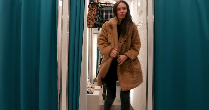 Woman dancing in a dressing room of a store in an artificial fur coat in interior boutique. Girl fun trying on outfit in checking room