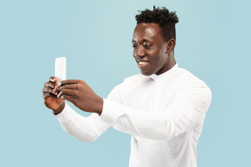 Let's tak a selfie. Young african-american man with smartphone. isolated on blue studio background, facial expression. Beautiful male half-lenght portrait. Concept of human emotions, facial expression