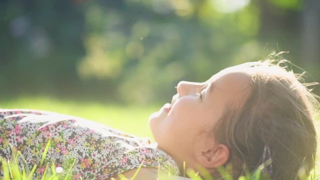 Portrait of a Happy smiling little girl lying on green grass, enjoying nature. Cute child outdoors. Healthy carefree kid playing outside in summer park or on backyard. Happiness. 4K slow motion 