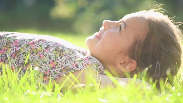 Portrait of a Happy smiling little girl lying on green grass, enjoying nature. Cute child outdoors. Healthy carefree kid playing outside in summer park or on backyard. Happiness. 4K slow motion 