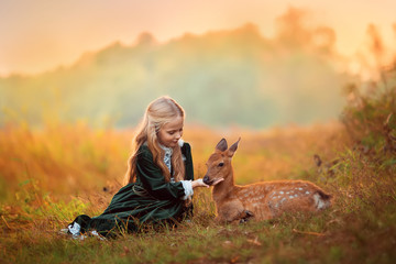 A cute little blonde girl in a green vintage dress sits next to a small sika deer around them...
