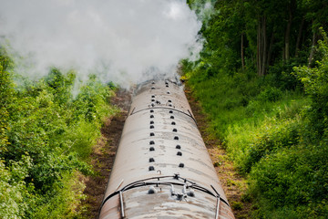 Railway bridge view of a famous, British steam locomotive seen pulling a number of passenger cars along a single track. Clouds of water seam is seen