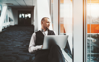 Fototapeta premium A confident serious prosperous caucasian man entrepreneur is sitting with a laptop on a white bench in a passageway of a modern business office skyscraper and pensively looking outside the window