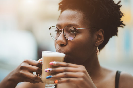 A close-up portrait of a young charming African female in spectacles outdoors holding a glass of delicious coffee latte; cute black girl in glasses and with nail art is drinking cocoa in a street cafe