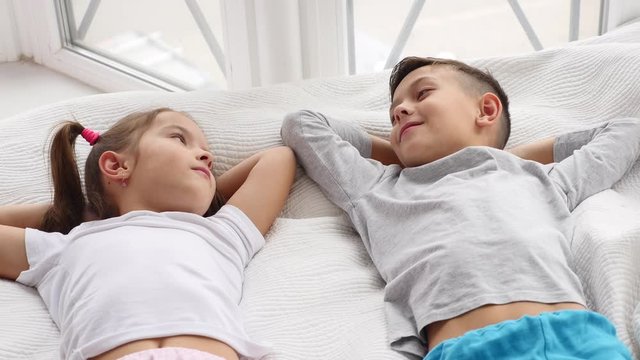 Top view on two beautiful siblings lying on a bed together, smiling and talking while having a rest.