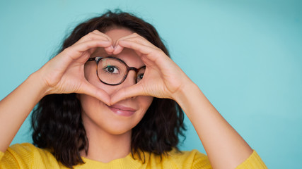 woman in glasses on a turquoise background makes a heart shape with her hands, a symbol of love - obrazy, fototapety, plakaty