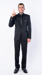 Full body shot of happy young bearded Persian businessman in suit pointing up