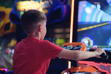 Cute European boy in red t-shirt playing car racing on slot-machine. He enjoying his spare time. Back view.