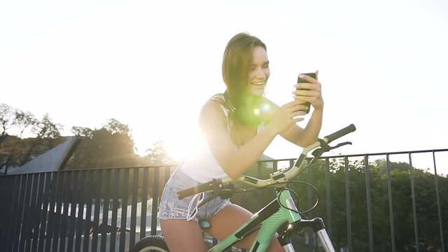 Portrait of attractive smiling girl in with long hair sits on bicycle and something watching in smartphone app in summer day at city