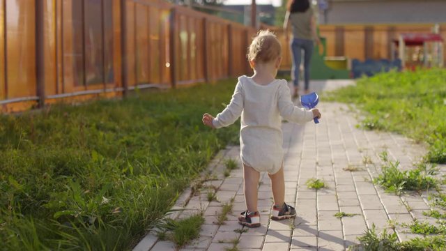 Full rear shot of blond toddler in bodysuit and sandals walking on paved footpath in private garden of suburban home, and woman and small dog far away