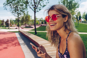 the student in sunglasses laughs and holds the phone in her hand. She is sitting in a park on a bench