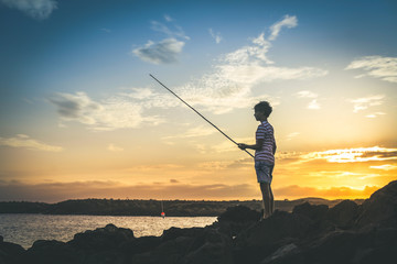 Young fisherman with fishing pole in hand looking the sea on a colorful sunset. Sailor boy with striped t-shirt enjoying a summer evening on the rocks. Lifestyle outdoors fun and carefree concept