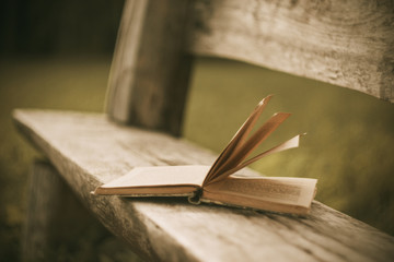 A forgotten open old book lies on a wooden bench cracked with time in the Park in the evening.