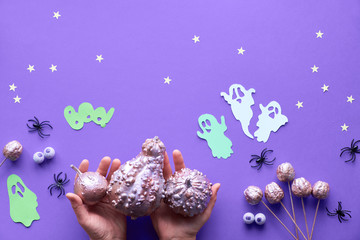 Creative Halloween flat lay on purple paper background with paper ghosts, spiders, stars and chocolate candy eyes. Hands holding gilded pink pumpkins.