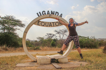 North-South Boundary Mark - Equator in Queen Elizabeth National Park in Uganda and a joyful Caucasian European tourist girl jumping.