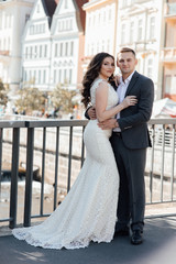 Fototapeta premium Handsome guy in suit and girl in white dress with a train on the bridge. A love story. Young romantic couple standing and hugging on a sunny day.