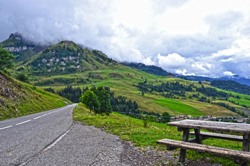 Fototapeta na wymiar La Clusaz, France - August 9th 2017 : Mountain landscape at 10 km of Albertville city. Focus on a road (with a picnic table) and wonderful green mountains in background, very cloudy.