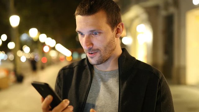 Excited adult man checking smart phone online content standing in the street in the night 