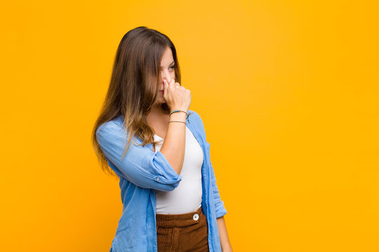 young pretty woman feeling disgusted, holding nose to avoid smelling a foul and unpleasant stench against orange wall