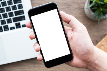Hand holding black smartphone with blank mock up screen over wood office desk table with laptop computer. Top view with copy space, flat lay.
