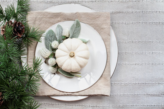 Thanksgiving Day or Christmas place setting with white plates, mini white pumpkins, Lamb's Ears leaves, flowers and pine branch over grey table runner.