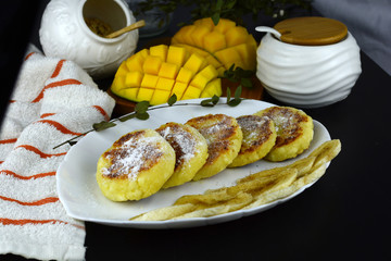 Homemade curd fritters, syrniki on a white plate. Next mango and a bowl of sugar.